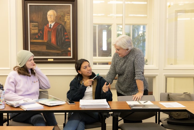 Two students are sitting at a desk, chatting with a School of Education professor in class. 