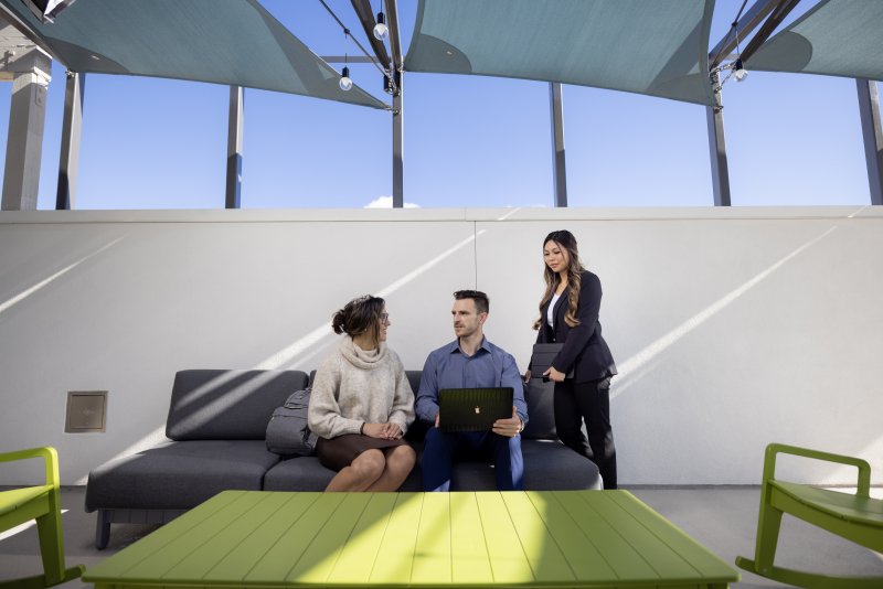 Three graduate program students are sitting in an outdoor venue. One has their laptop open and they are talking amongst eachother.