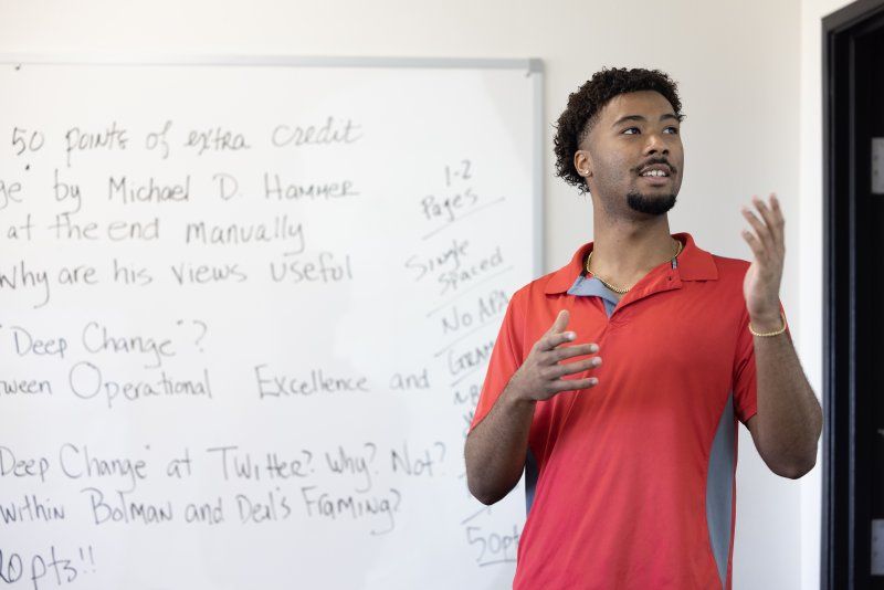 A student in a red shirt gives a presentation in front of a white board