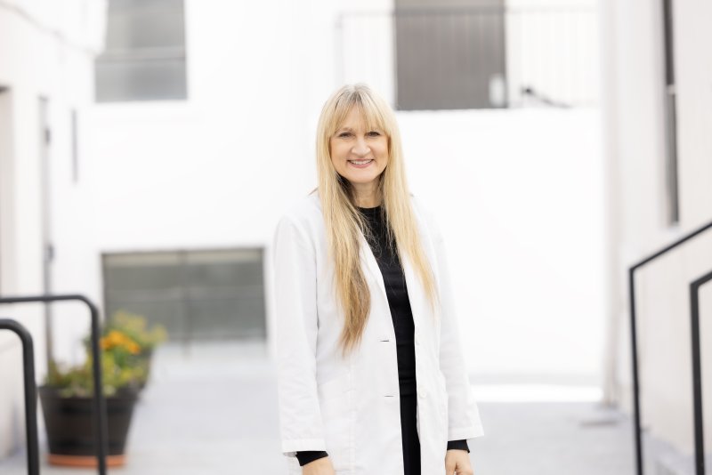 A female nurse with blonde hair is smiling. She is wearing a white coat and black scrubs and has a stethoscope around her neck. 