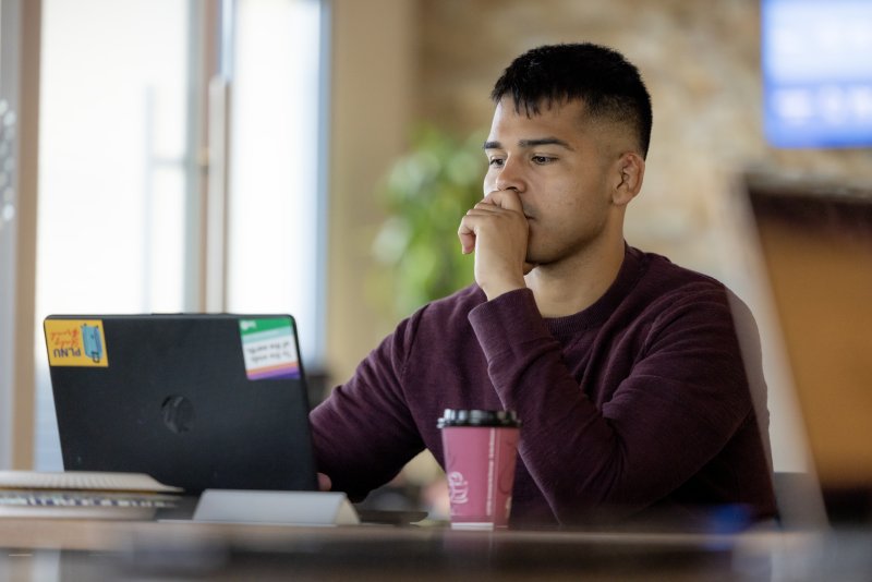 A male student is sitting at a desk and working on a graphic design project on his laptop.