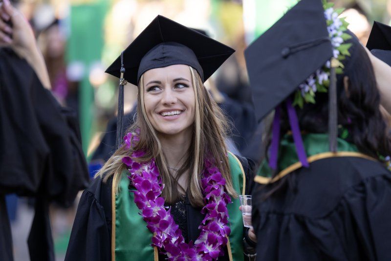 A female student with brown hair is smiling at her PLNU graduation ceremony. She is wearing a black cap and gown, as well as a purple lei.