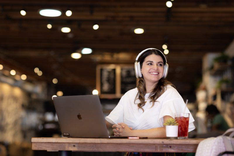 A woman is smiling and sitting in a coffee shop. She is wearing headphones and has an open laptop in front of her.