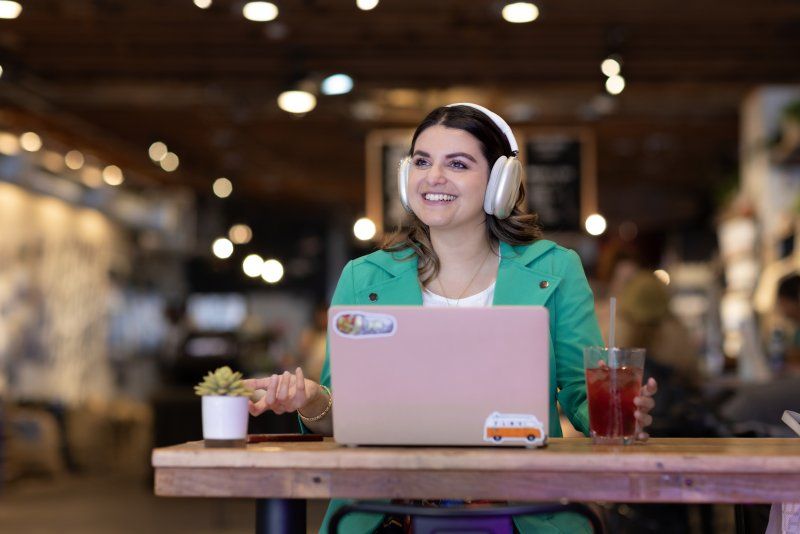 A woman in a green jacket is sitting in a coffee shop, smiling. She is wearing headphones and has an open laptop and beverage on the table in front of her.