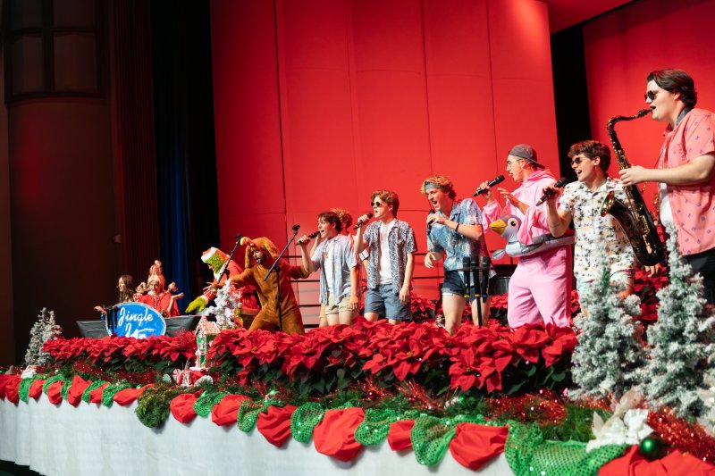 several college students singing into microphones, wearing christmas outfits and beach outfits. The backdrop is red and the stage is lined with red poinsettia flowers.