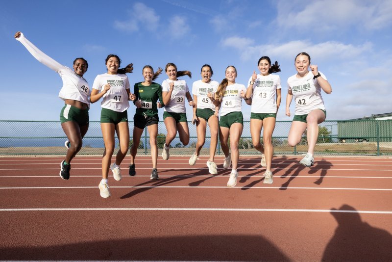 Female PLNU athletes all jump in celebration at a track meet on the university track with the Pacific Ocean in the background.