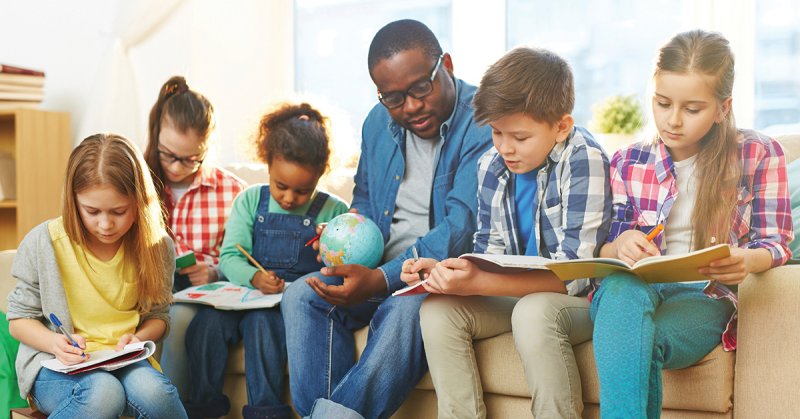 A teacher sits on the couch with five children and teaches them about geography.