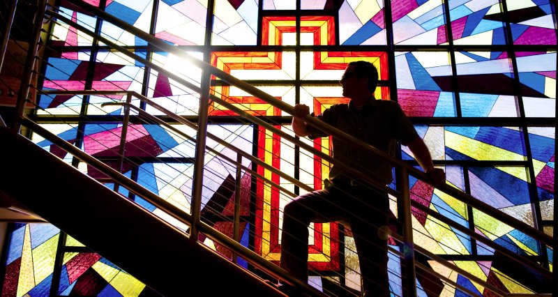 Man climbs staircase in shadow, backlit by sun coming through giant stained glass window of a cross