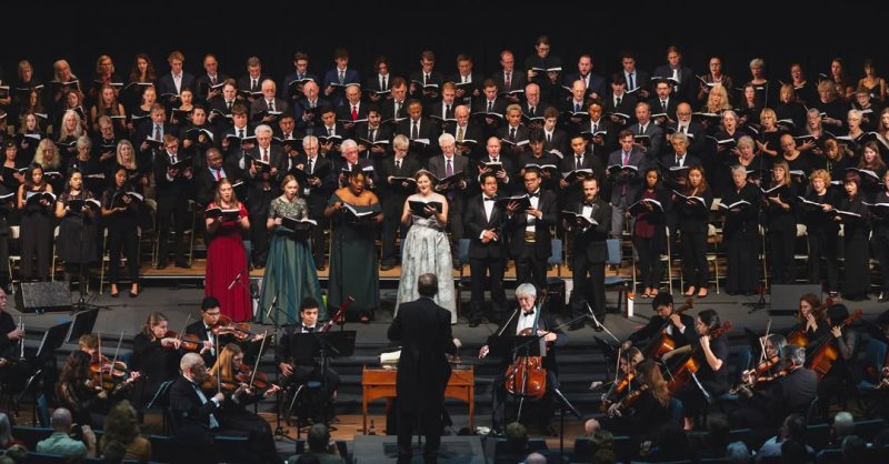 large choir with 7 featured singers in front. The conductor and orchestra are below the choir. 