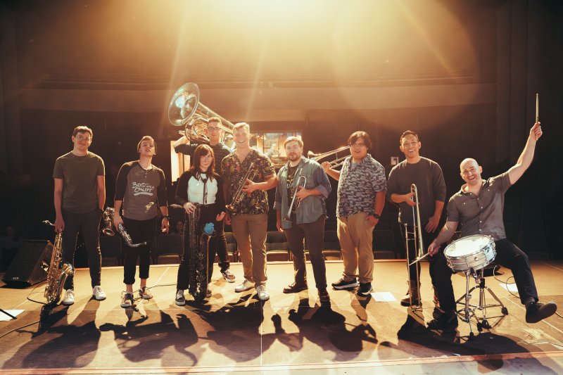 9 muscians on a performance stage holding various brass and percussion instruments