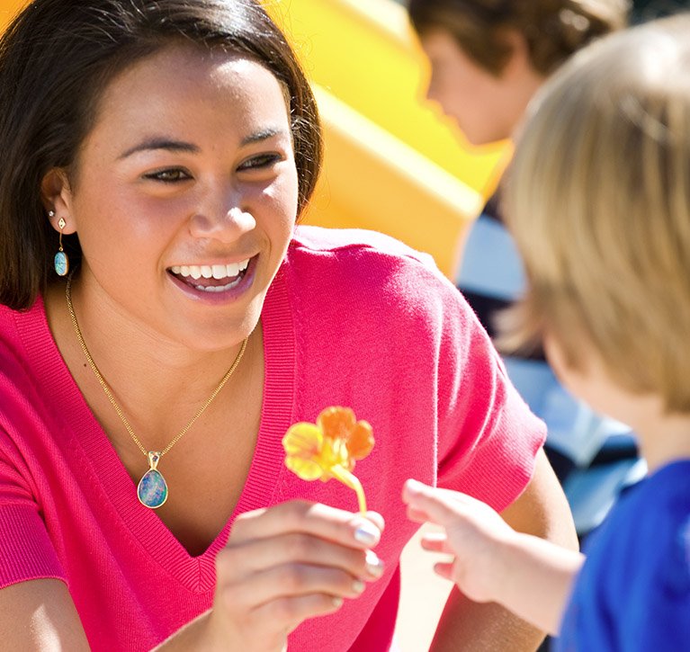 A female ECLC student worker hands a small orange flower to a child in the playground