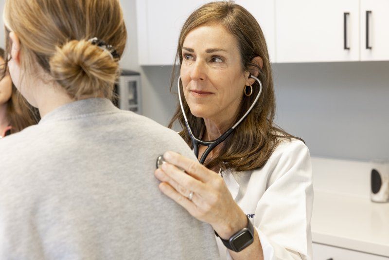 A female nurse with brown hair is holding a stethoscope to her patient's back.