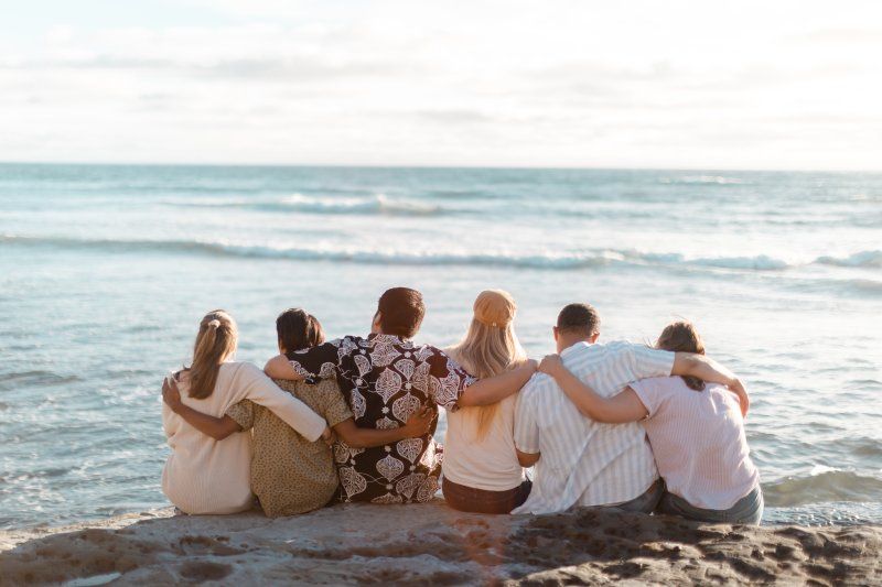 Six friends sit on the sand, facing the ocean in front of them. They all have their arms wrapped around each other. 