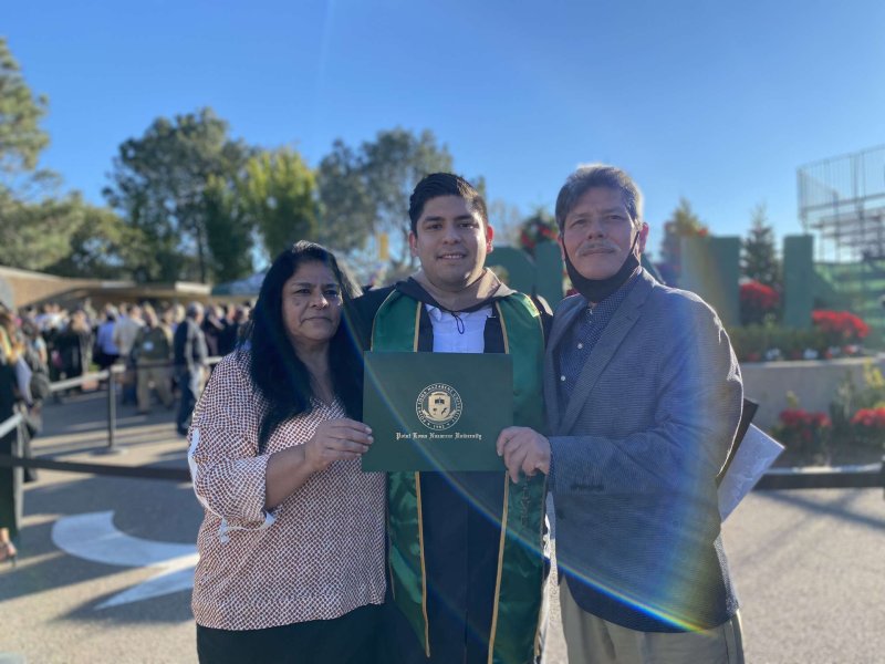 Erick is posing after graduation holding up his diploma with his parents to his left and right. 