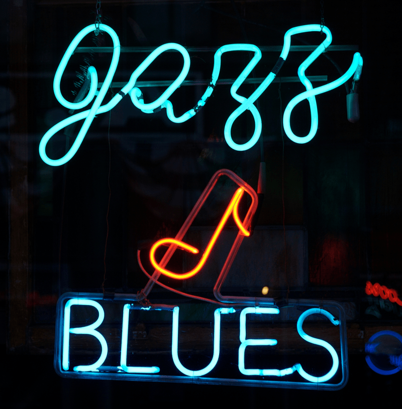 Image of the words 'jazz' and 'blues' lit up at night with a neon blue sign with a music note also lit up with an orange neon sign.