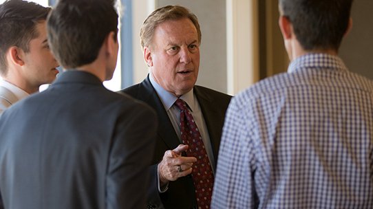 Male professor speaks with a group of men during a networking event