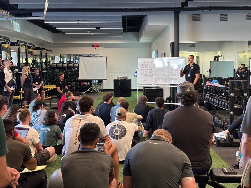 A group of students and professionals sit on turf flooring inside a modern training facility, listening to a speaker standing beside a whiteboard filled with notes. Weight racks, barbells, and gym equipment line the room as the presenter gestures while leading an instructional session.