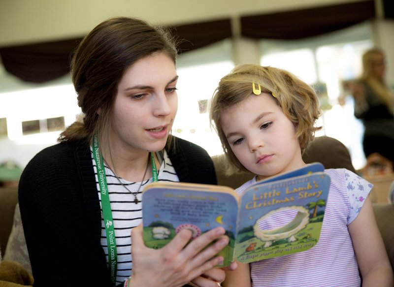 A female college student reads a children's book to a young girl inside
