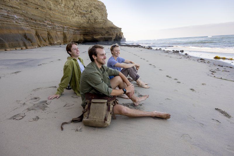 Three male students sit together on the beach, smiling and looking toward the ocean at the right. The two males on the left are wearing green shirts, and the one male student on the right is wearing a blue shirt.