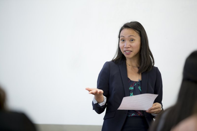 A woman in a black blazer is holding a piece of paper and speaking as she stands in front of a white board.