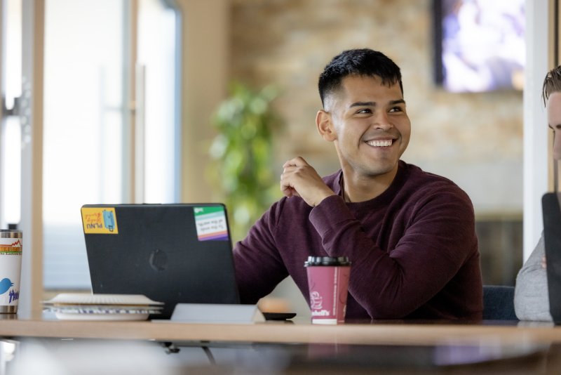 A male in a maroon sweater sits at a desk, smiling as he looks to his left. His laptop is open on the desk in front of him.