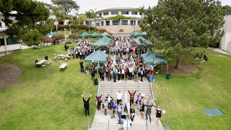 Large group of students, faculty, and research presenters gather outdoors on the PLNU campus during an academic research event.