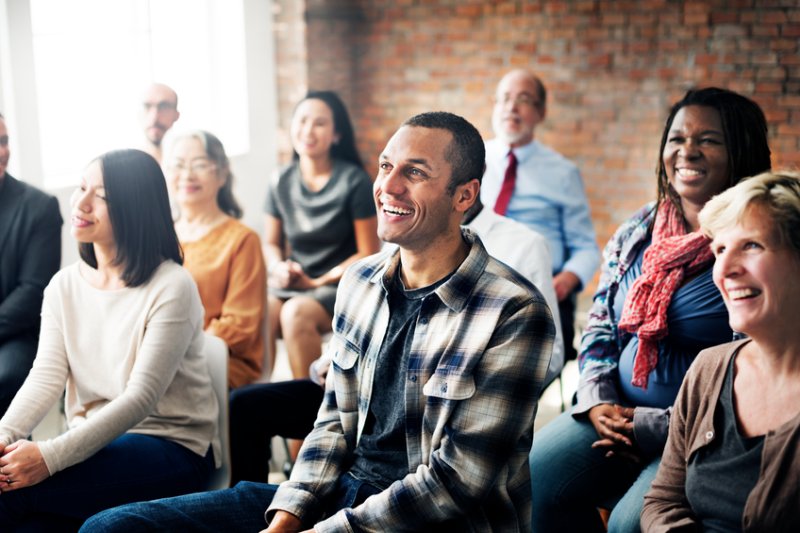 A diverse group of people from different backgrounds and age ranges sit in a brick building and listen to a speaker.