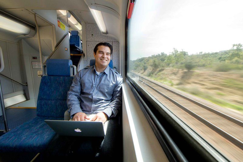 online graduate student sitting in a train with a laptop on his lap. he is looking outside of the train smiling