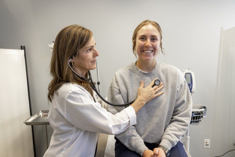 A female nurse with brown hair is holding a stethoscope to her patient's heart. The patient is a female with blonde hair. She is sitting on the exam table, smiling.