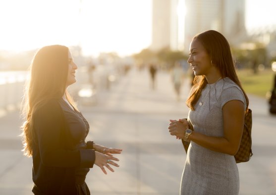 Two women stand outside, looking at each other while in conversation. The one on the left is in a black cardigan and the one on the left is in a grey dress.