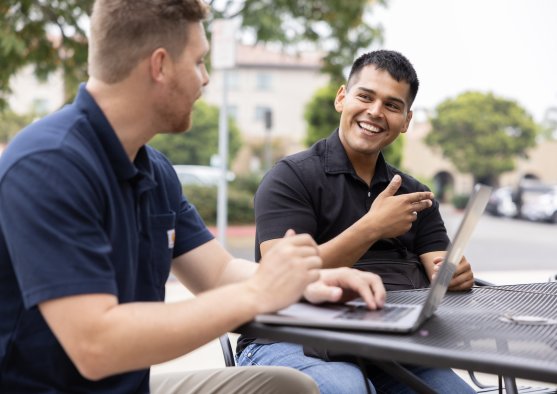 Two PLNU students work on laptops outside 