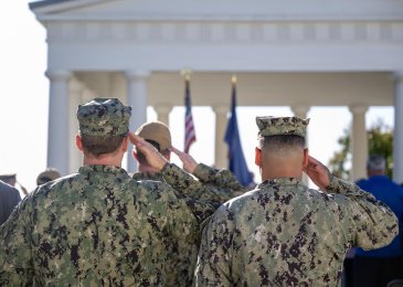 Uniformed military salute the American flag with the camera angled from behind the service members.
