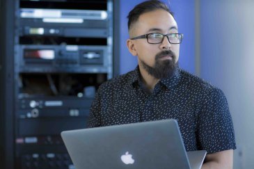 PLNU member working on a laptop in a technology lab with server equipment in the background.